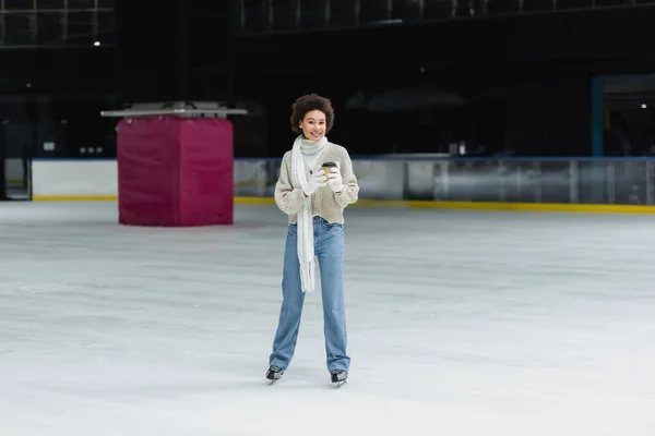 Allegro donna afro-americana in possesso di tazza di carta e guardando la fotocamera sulla pista di pattinaggio sul ghiaccio — Foto stock