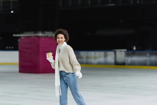 Sonriente mujer afroamericana en bufanda y guantes sosteniendo una taza de papel y mirando a la cámara en la pista de hielo — Stock Photo