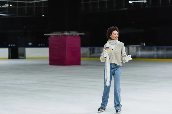 Young african american woman ice skating and holding coffee to go on rink — Stock Photo