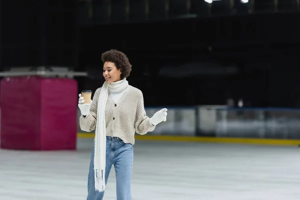 Positive african american woman in gloves and warm clothes holding coffee to go on ice rink — Stock Photo