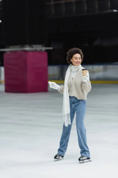 African american woman ice skating and looking at coffee to go on rink — Stock Photo