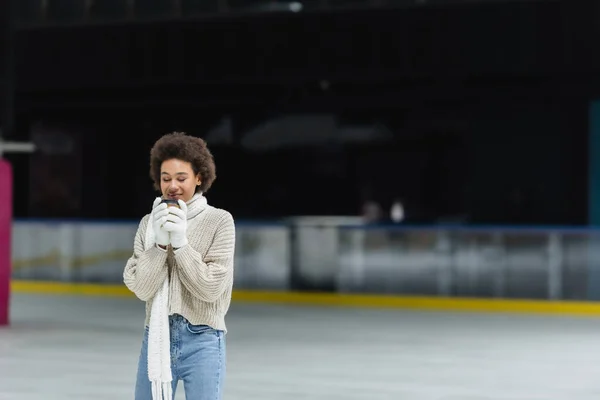 Joven mujer afroamericana en guantes y bufanda sosteniendo café para ir en pista de hielo - foto de stock