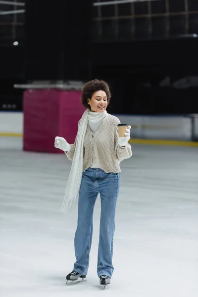 Smiling african american woman ice skating and looking at takeaway coffee in paper cup on ice rink — Stock Photo