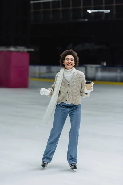 Mujer afroamericana positiva sosteniendo café para ir mientras patina sobre hielo y mirando la cámara en la pista - foto de stock