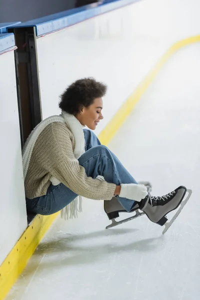 Vista lateral da mulher afro-americana positiva amarrando atacadores de patins de gelo na pista — Fotografia de Stock