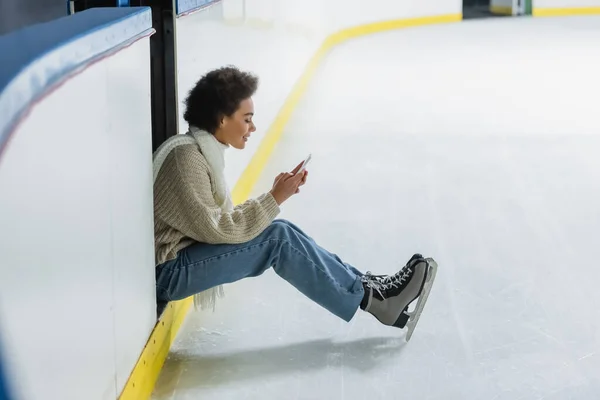Vista laterale della donna afroamericana in pattini da ghiaccio utilizzando smartphone sulla pista di pattinaggio — Foto stock