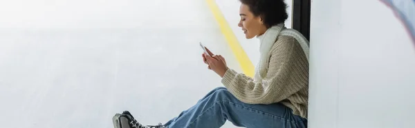 Vista lateral de la mujer afroamericana en suéter cálido con teléfono inteligente en pista de hielo, pancarta - foto de stock
