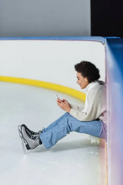 Vista laterale di allegra donna afroamericana in pattini da ghiaccio e sciarpa utilizzando smartphone sulla pista di pattinaggio — Foto stock