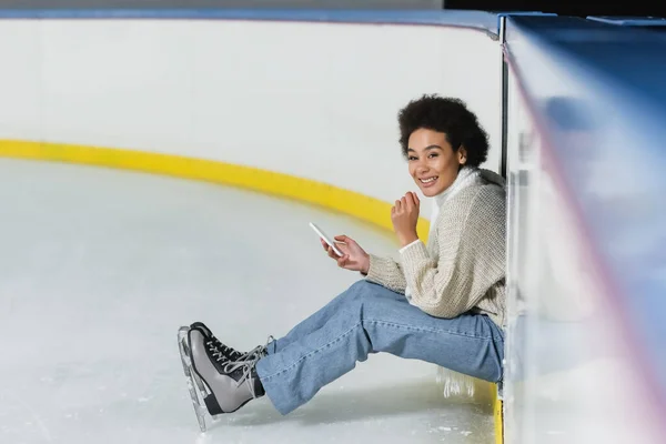 Mulher afro-americana positiva segurando smartphone e olhando para a câmera na pista de gelo — Fotografia de Stock