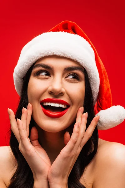 Porträt einer aufgeregten Frau mit Visage und Weihnachtsmütze, die Wangen berührt, isoliert auf Rot — Stockfoto