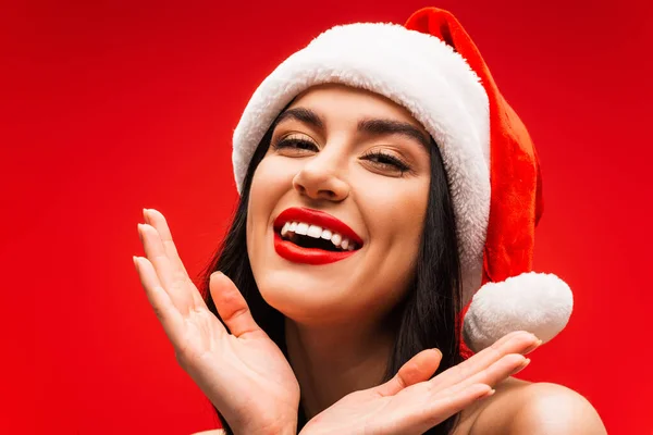 Portrait of positive young model in santa hat looking at camera isolated on red — Stock Photo