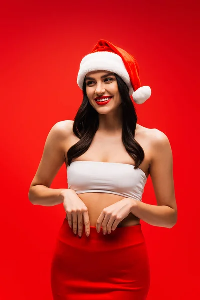 Pretty young woman in santa hat and top touching skirt isolated on red — Stock Photo