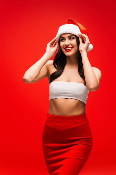 Pretty young woman in top and skirt touching santa hat isolated on red — Stock Photo