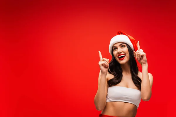 Cheerful brunette model in top and santa hat pointing with fingers isolated on red — Stock Photo