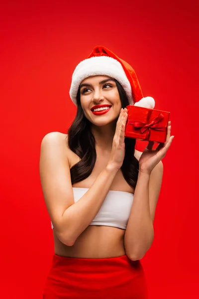 Cheerful woman in santa hat and top holding present isolated on red — Stock Photo