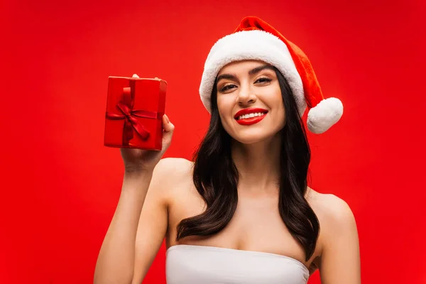 Positive brunette model in santa hat holding present isolated on red — Stock Photo