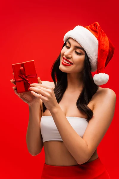 Mulher morena alegre em Papai Noel olhando caixa de presente isolado em vermelho — Fotografia de Stock