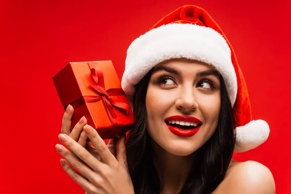 Porträt einer jungen Frau mit Make-up und Weihnachtsmannmütze, die ein Geschenk in Ohrennähe hält, isoliert auf Rot — Stockfoto