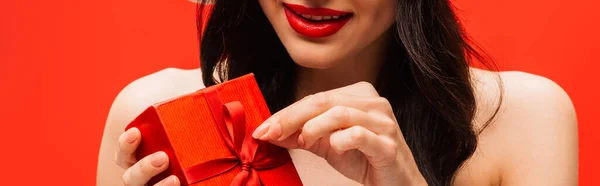 Vista recortada de mujer joven con maquillaje y hombros desnudos tocando arco en regalo aislado en rojo - foto de stock
