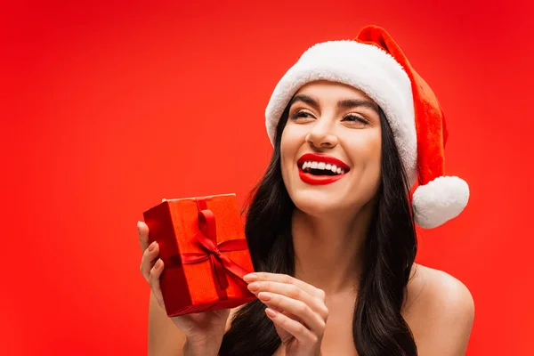 Donna sorridente in cappello di Babbo Natale che tiene la scatola regalo e distoglie lo sguardo isolato sul rosso — Foto stock