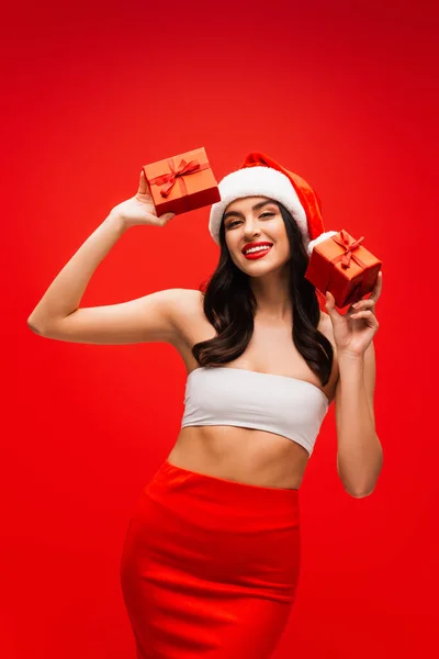 Sexy woman in santa hat holding gifts and looking at camera isolated on red — Stock Photo