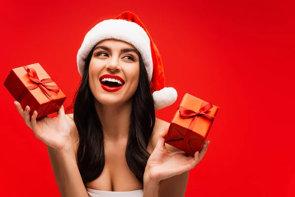 Портрет улыбающейся модели с визажем и шляпой Санты холдинг подарки изолированы на красный — стоковое фото