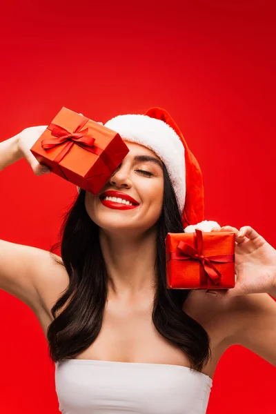Mujer sonriente en la parte superior y sombrero de santa sonriendo y sosteniendo regalos aislados en rojo - foto de stock