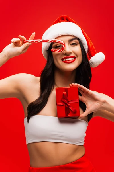 Lächelndes Model mit Weihnachtsmütze, Weihnachtslutscher und Geschenk isoliert auf Rot — Stockfoto