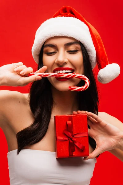 Brünette Frau mit Zylinder und Weihnachtsmütze beißt Weihnachtslutscher und hält Geschenk isoliert auf rot — Stockfoto