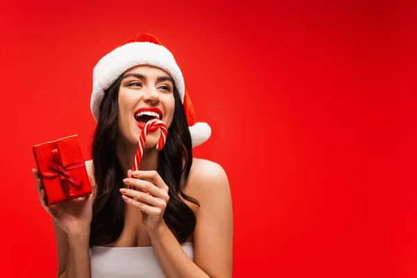 Excited woman in top and santa hat holding striped lollipop and present isolated on red — Stock Photo