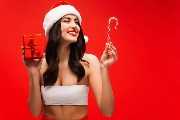 Smiling model in top and santa hat holding striped lollipop and gift box isolated on red — Stock Photo
