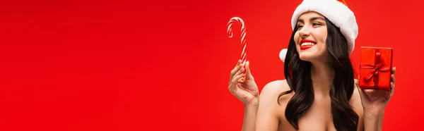 Hübsches Modell mit Weihnachtsmütze, Geschenk und gestreiftem Lutscher, isoliert auf rot, Banner — Stockfoto