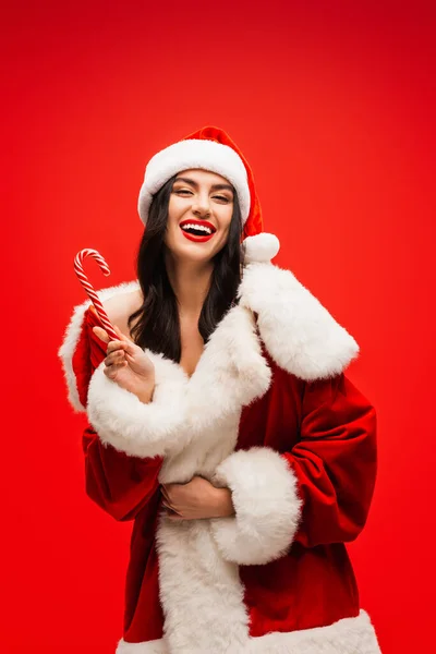 Cheerful young model in santa costume holding striped lollipop isolated on red — Stock Photo