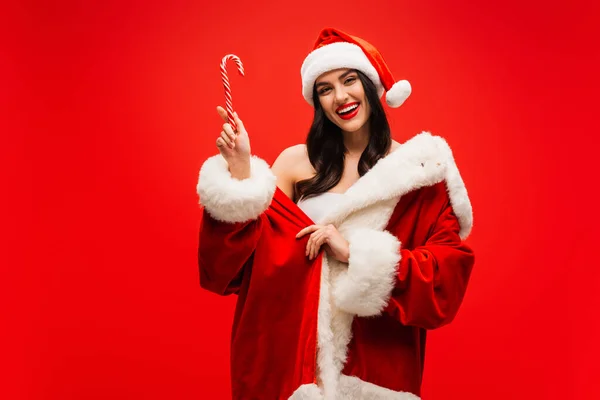 Cheerful brunette woman in santa costume holding striped lollipop isolated on red — Stock Photo