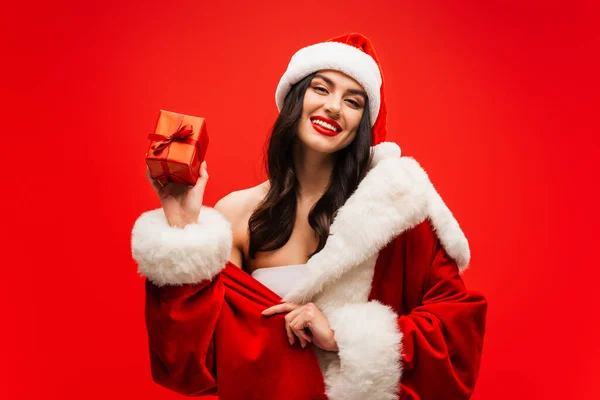 Modello positivo in costume da Babbo Natale in possesso presente e guardando la fotocamera isolata sul rosso — Foto stock