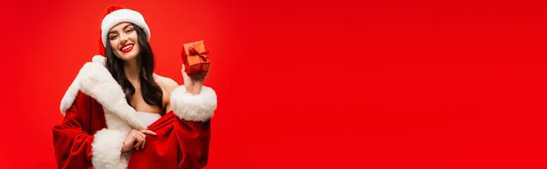 Pretty woman in santa costume holding gift box and smiling isolated on red, banner — Stock Photo