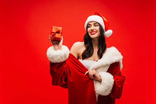 Pretty brunette woman in santa costume looking at present with ribbon isolated on red — Stock Photo