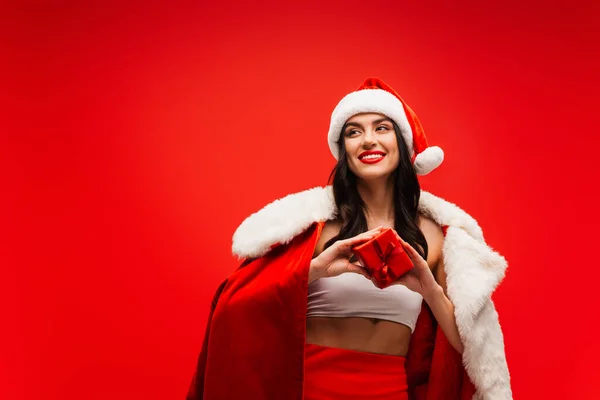 Pretty young woman in santa costume and hat holding gift box isolated on red — Stock Photo