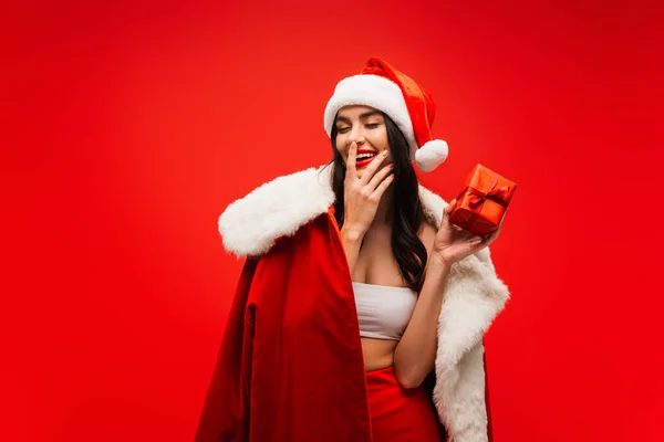 Jeune femme en costume de Père Noël et chapeau tenant présent et touchant visage isolé sur rouge — Photo de stock