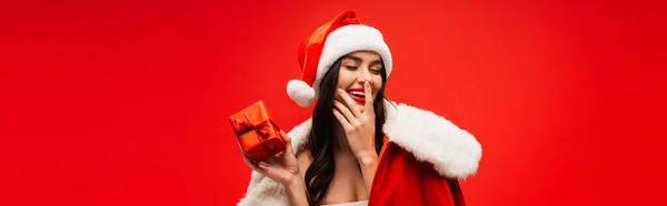 Pretty brunette model in santa costume and hat holding gift and touching face isolated on red, banner — Stock Photo