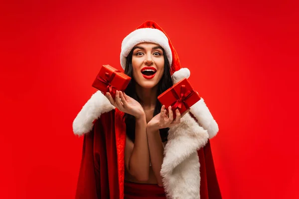 Mujer excitada en sombrero de santa y traje sosteniendo regalos aislados en rojo - foto de stock