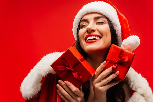 Femme heureuse dans le chapeau santa tenant des boîtes-cadeaux et les yeux fermés isolés sur rouge — Photo de stock