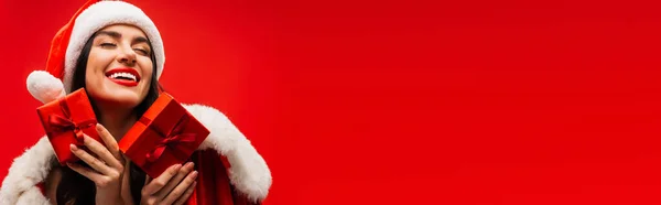 Femme agréable en costume de Père Noël tenant des boîtes-cadeaux isolées sur rouge avec espace de copie, bannière — Photo de stock