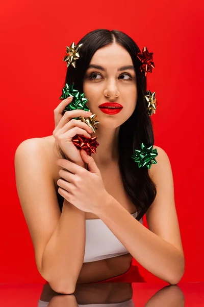 Jeune femme avec maquillage tenant des noeuds cadeaux près du miroir isolé sur rouge — Photo de stock