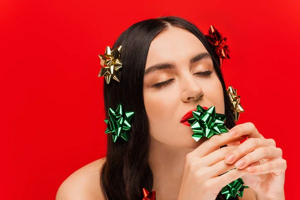 Young brunette woman with visage holding gift bows near lips isolated on red — Stock Photo