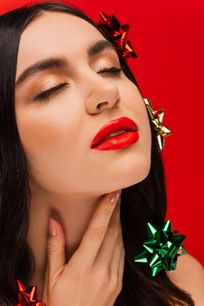 Vue rapprochée de la femme brune avec maquillage et noeuds cadeaux sur les cheveux touchant le cou isolé sur rouge — Photo de stock