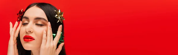 Modelo jovem com arcos de presente no cabelo tocando rosto isolado no vermelho com espaço de cópia, banner — Fotografia de Stock