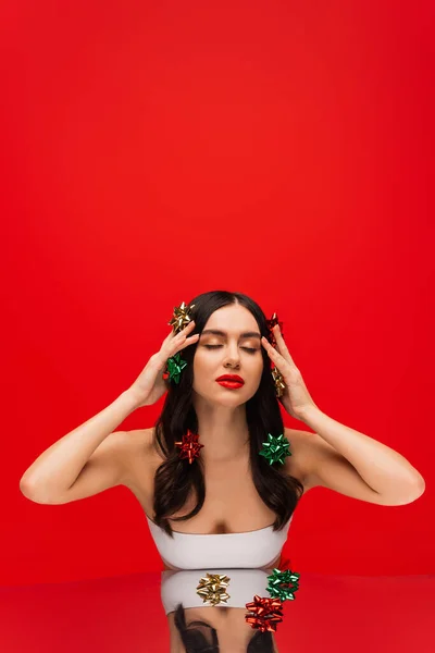 Modello giovane con fiocchi regalo sui capelli in posa vicino alla superficie riflettente isolata sul rosso — Foto stock