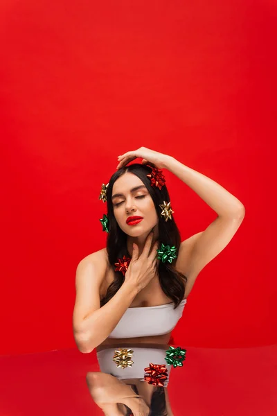 Donna bruna con fiocchi regalo sui capelli che toccano il collo vicino allo specchio isolato sul rosso — Foto stock
