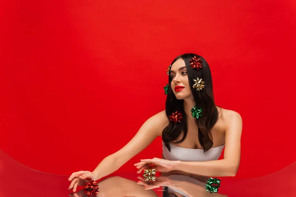 Giovane donna con spalle nude e fiocchi regalo sui capelli posa vicino specchio isolato su rosso — Foto stock